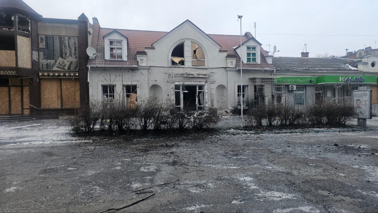 Фасад одноповерхової комерційної будівлі зі значними пошкодженнями після обстрілу: вибиті вікна, обгорілі стіни та сліди прильотів на фоні порожньої площі.