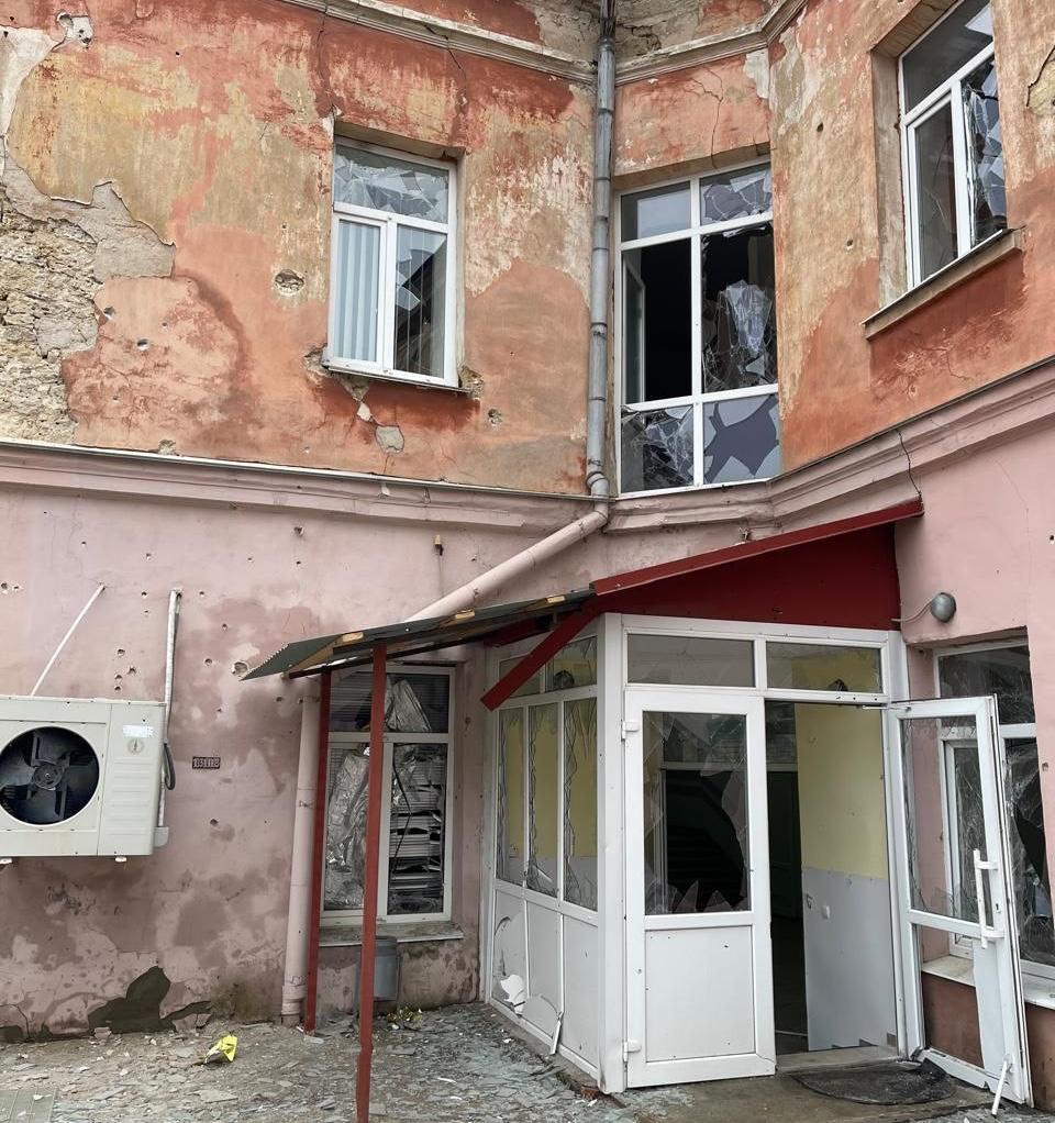 Пошкоджена двоповерхова будівля з посіченими стінами, розбитими вікнами та зруйнованою скляною вхідною групою.