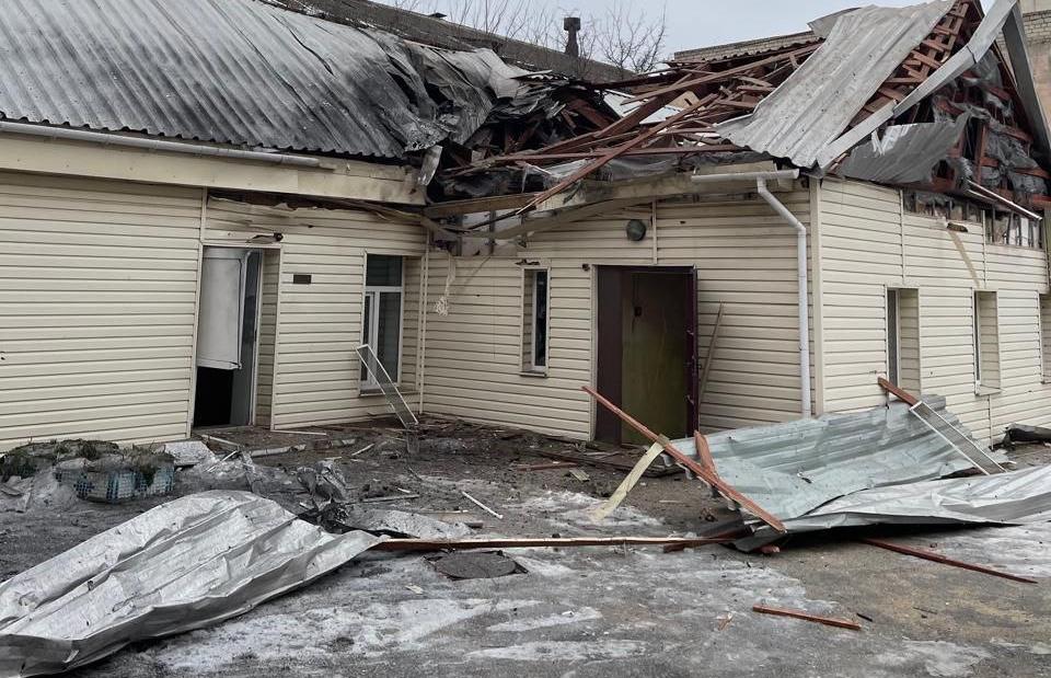 Одноповерхова будівля, обшита світлим сайдингом, із серйозними руйнуваннями даху: покрівля розтрощена, балки перекриття оголені, а листи металопрофілю та будівельне сміття розкидані на землі перед входом.