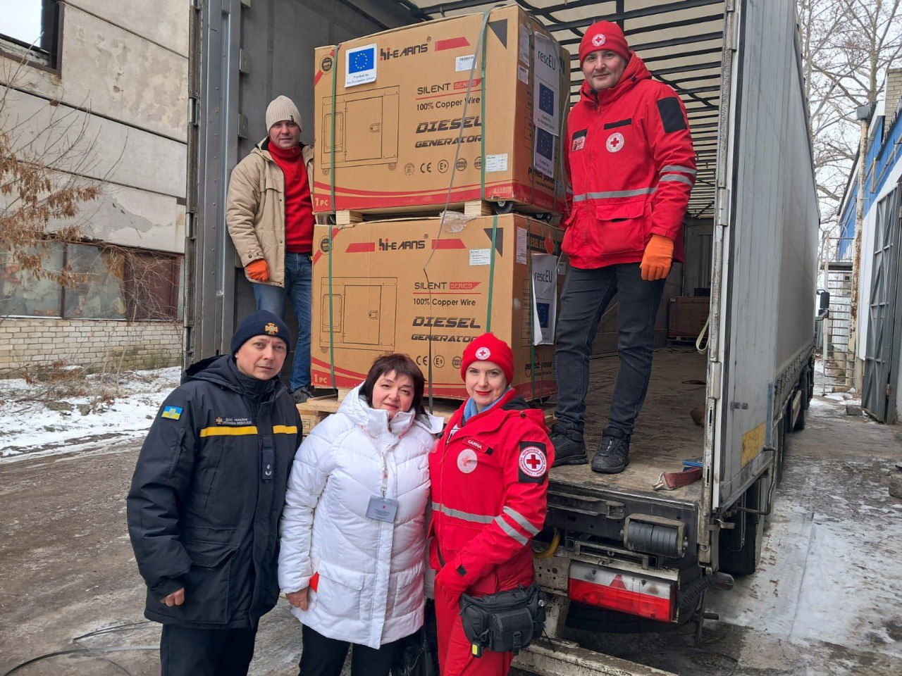 Представники гуманітарних служб та рятувальники стоять біля вантажівки з великими коробками, у яких доставлено дизельні генератори для забезпечення енергетичних потреб громади.