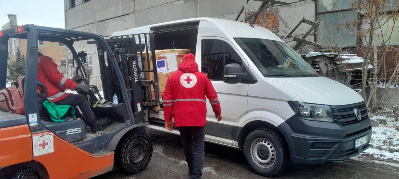 Навантажувач переміщує великий ящик із гуманітарною допомогою до білого мікроавтобуса під наглядом працівника в червоному форменому одязі з емблемою Червоного Хреста.