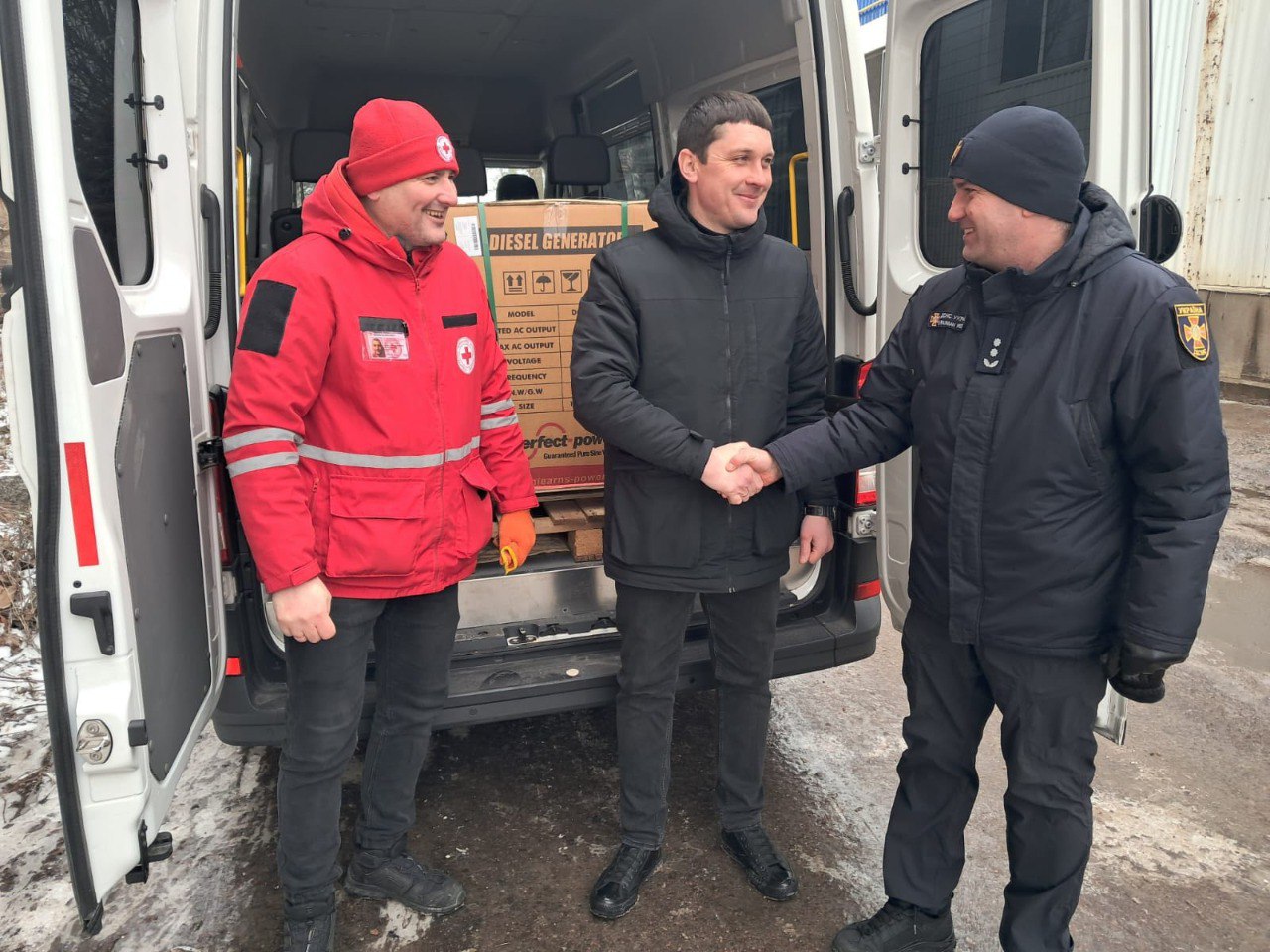 Представник ДСНС та працівник Червоного Хреста тиснуть руки чоловікові на фоні відчиненого мікроавтобуса, у якому знаходиться дизельний генератор у великій картонній коробці.