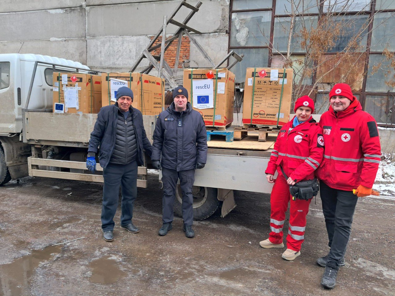 Представники рятувальних та гуманітарних служб стоять поруч із вантажним автомобілем, на платформі якого розміщено кілька великих коробок із дизельними генераторами, маркованими символікою ЄС та гуманітарних програм.