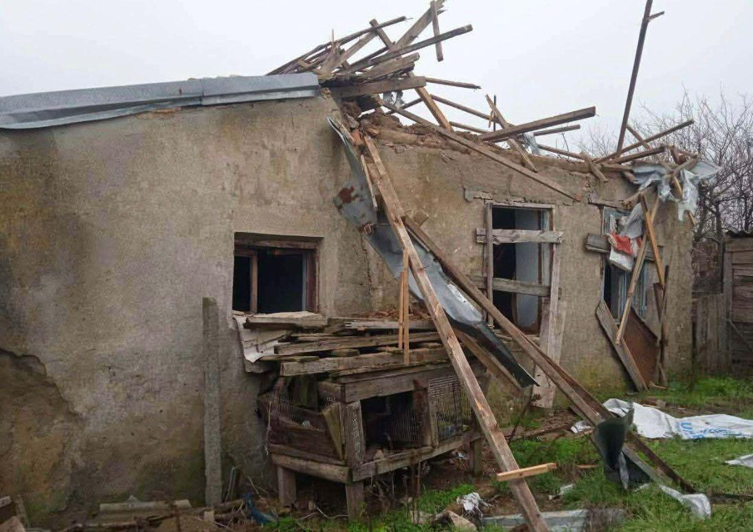 Напівзруйнована невелика господарська споруда або приватний будинок із розтрощеним дахом, де балки перекриття впали на землю, а вікна та частина стіни мають серйозні пошкодження.