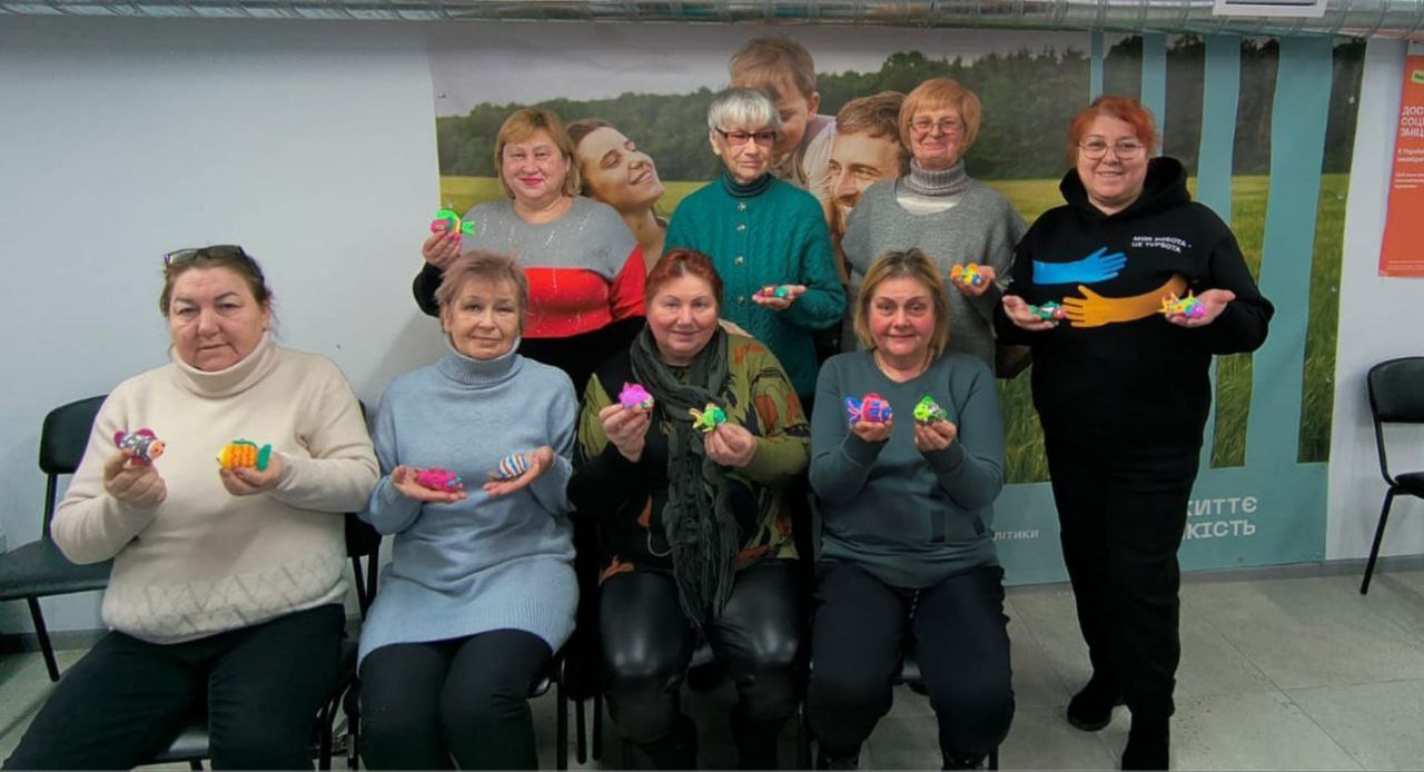 Група жінок у світлому приміщенні позує для спільного фото, тримаючи в руках яскраві різнокольорові вироби ручної роботи у формі рибок. На фоні видно плакат із зображенням сім’ї та текст про соціальну підтримку.