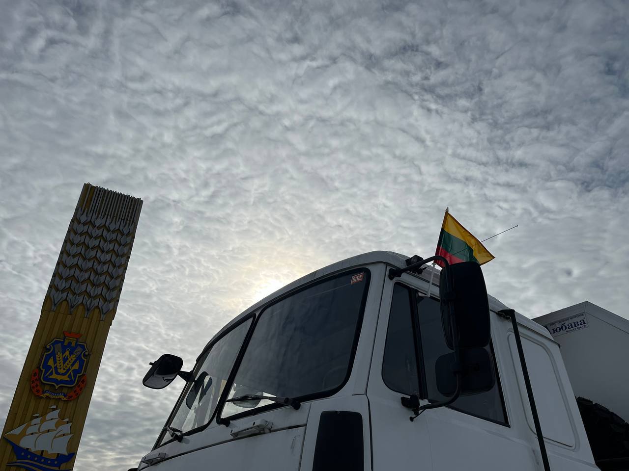 Біла вантажівка з невеликим прапором Литви на дзеркалі заднього виду стоїть на фоні стели Херсонської області під хмарним небом.