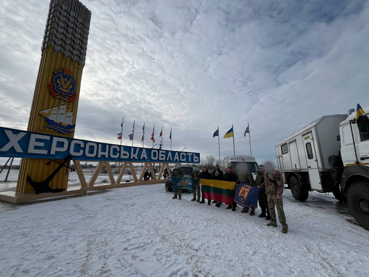 Група людей стоїть на засніженій дорозі біля в’їзного знаку «Херсонська область». Вони тримають прапори, серед яких прапор Литви та символіка підрозділів. Поруч припарковані вантажівка та позашляховики.