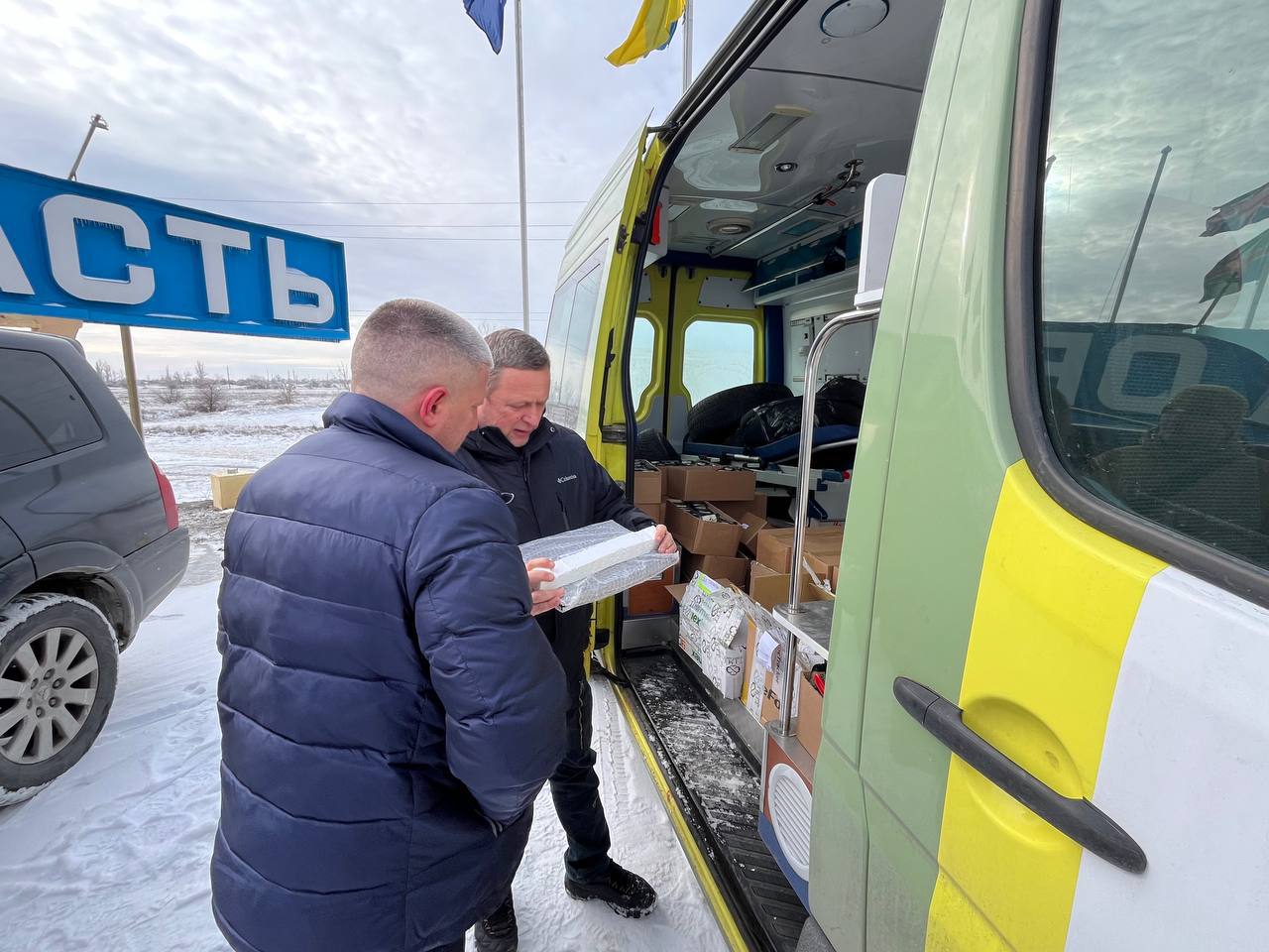 Двоє чоловіків розвантажують гуманітарну допомогу з мікроавтобуса на засніженому узбіччі. Один із них тримає пакунок у захисній плівці, а всередині автомобіля видно багато картонних коробок. На фоні височіє в’їзний знак Херсонської області.