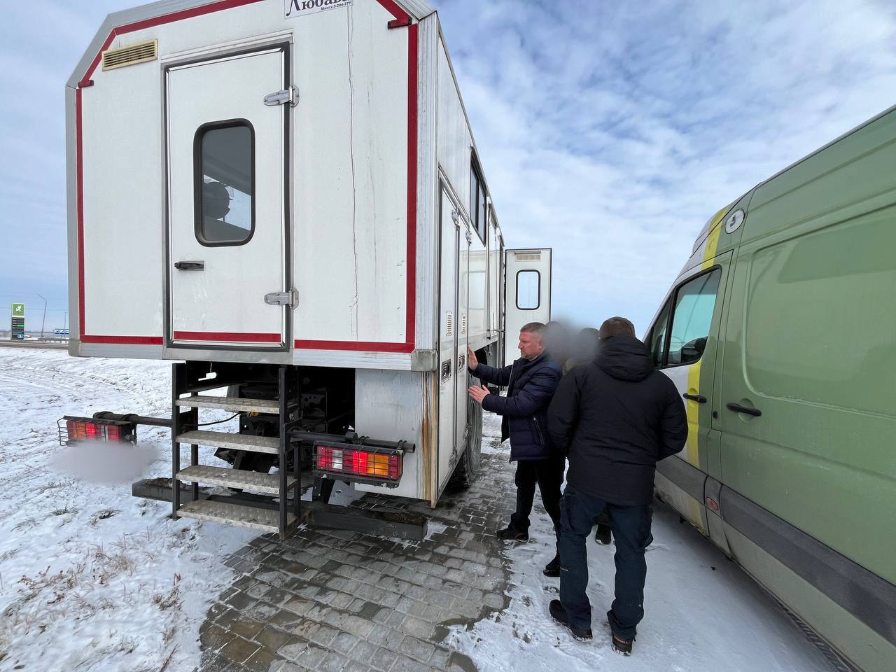 Двоє чоловіків стоять на засніженому узбіччі поруч із задньою частиною великого білого спеціалізованого фургона та зеленим мікроавтобусом. Вони оглядають двері фургона, обладнаного сходами.