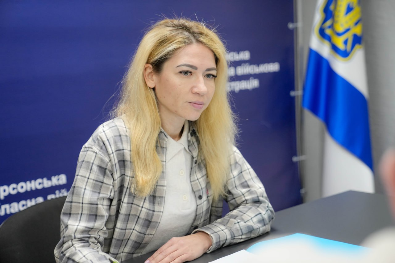 Жінка у світлій сорочці в клітинку сидить за столом під час офіційного заходу. На задньому плані видно синій банер із символікою Херсонської обласної військової адміністрації та Державний Прапор України.