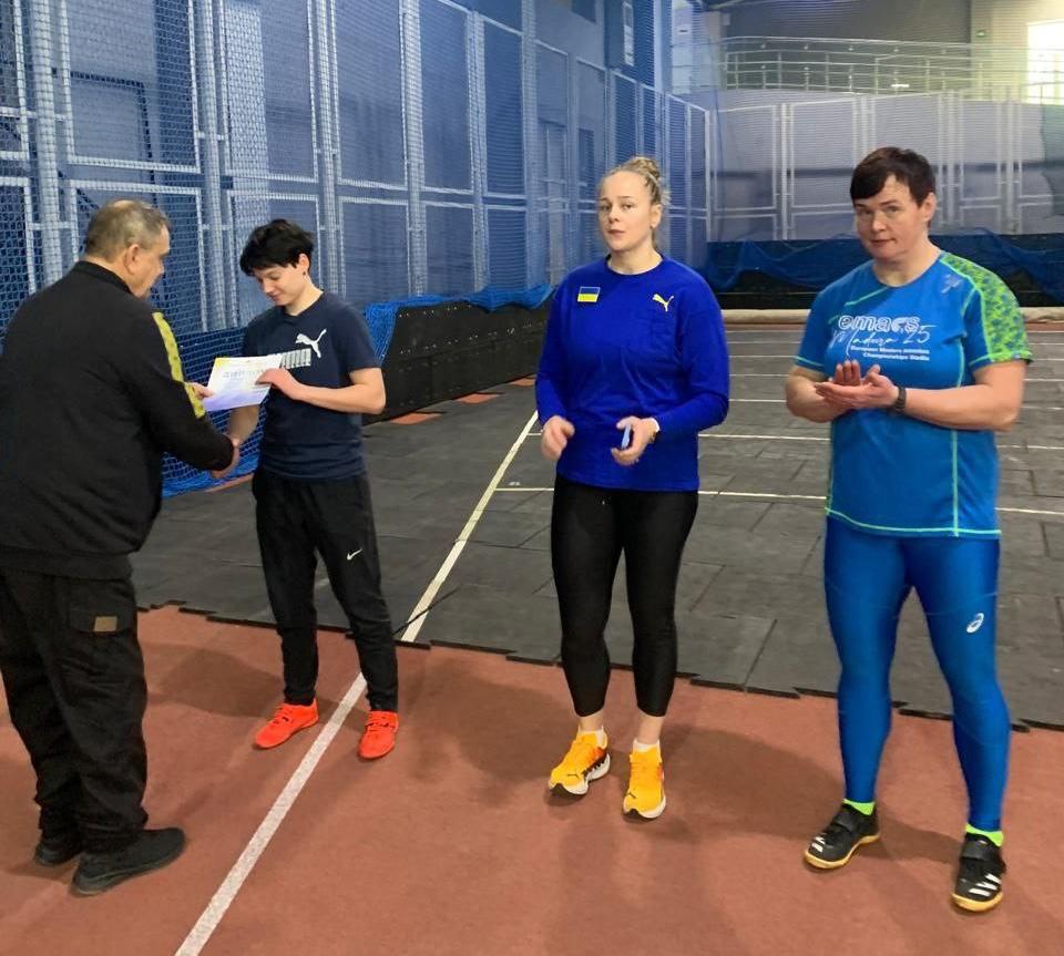 У спортивному залі чоловік вручає диплом підлітку в спортивному одязі. Поруч стоять двоє дорослих спортсменів: жінка у синьому лонгсліві з українською символікою та жінка у синьо-зеленій спортивній формі, яка аплодує.