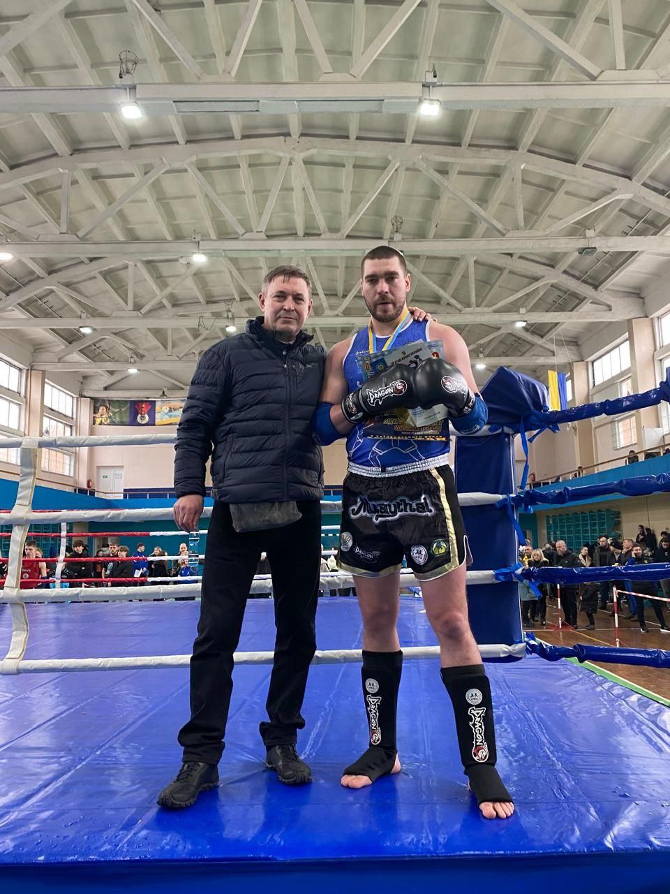 На боксерському рингу стоять двоє чоловіків: спортсмен у синій формі, екіпіруванні для тайського боксу та з медаллю на шиї, і чоловік у темній куртці, який поклав йому руку на плече. На задньому плані видно спортивний зал та інших глядачів.