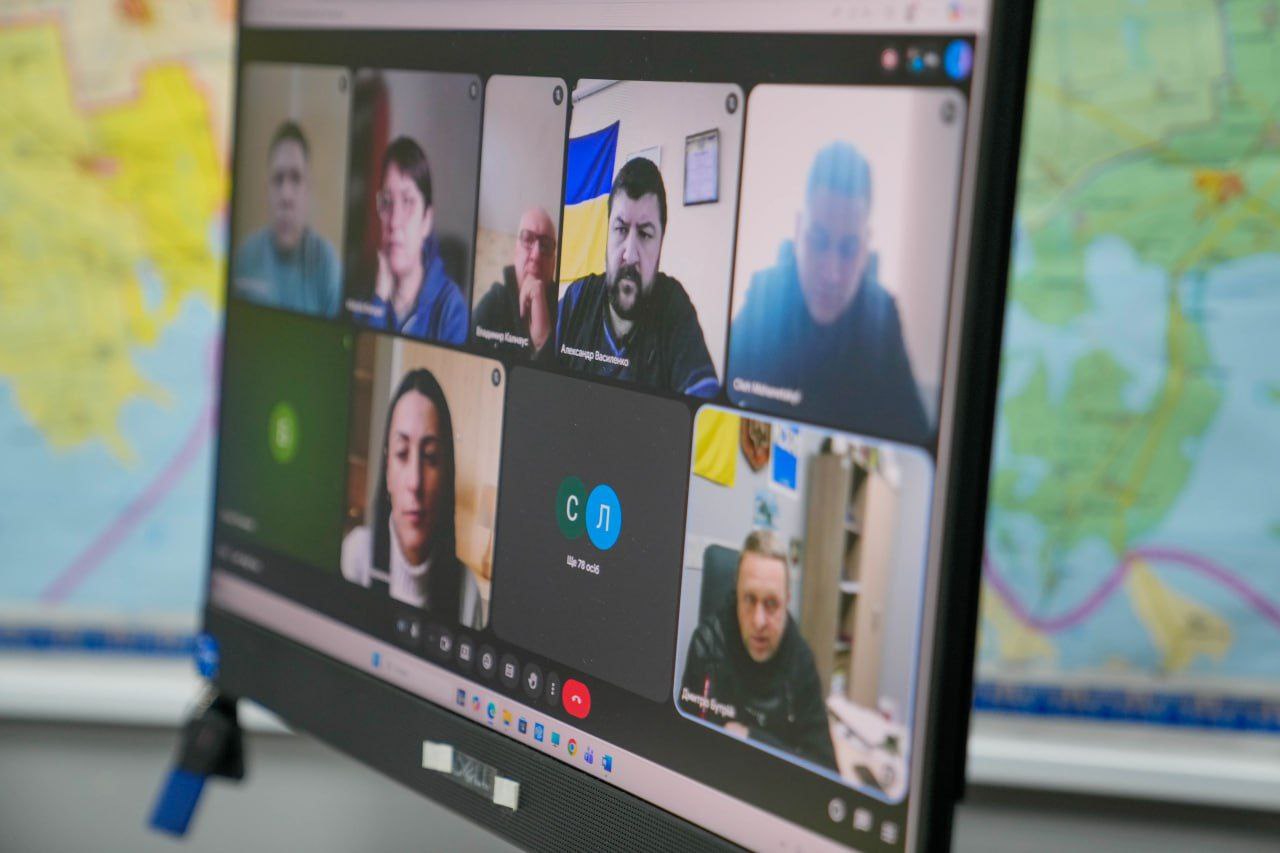 Екран монітора, на якому відображено вікно онлайн-конференції з кількома учасниками. На задньому плані за монітором видно частину географічної карти.