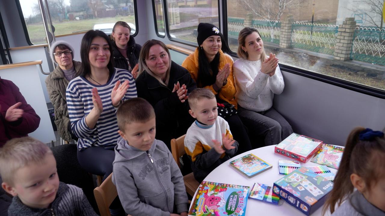 Група жінок та дітей сидить за столом усередині спеціально облаштованого автобуса. Вони посміхаються та аплодують, а на столі перед ними лежать дитячі книжки, розмальовки та набори для творчості.