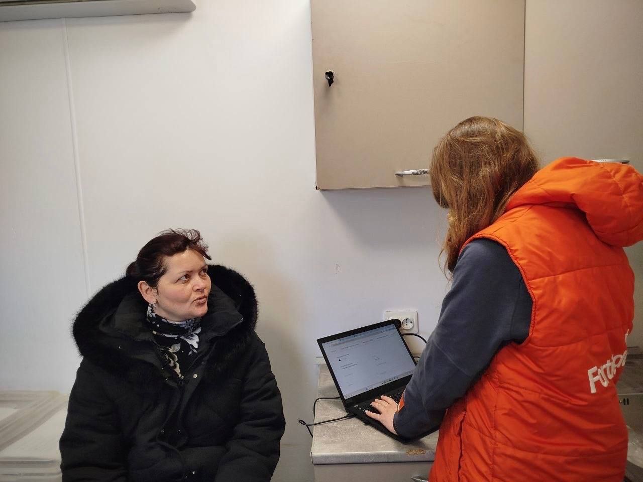 Медична працівниця у помаранчевому жилеті вносить дані до ноутбука під час консультації пацієнтки в мобільному кабінеті.