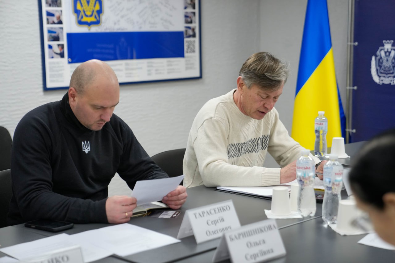 Двоє чоловіків сидять за робочим столом під час наради. Один із них, у чорному світшоті з гербом України, вивчає документи, а інший, у світлому светрі з орнаментом, робить нотатки. На фоні видно Державний Прапор України та інформаційний стенд.