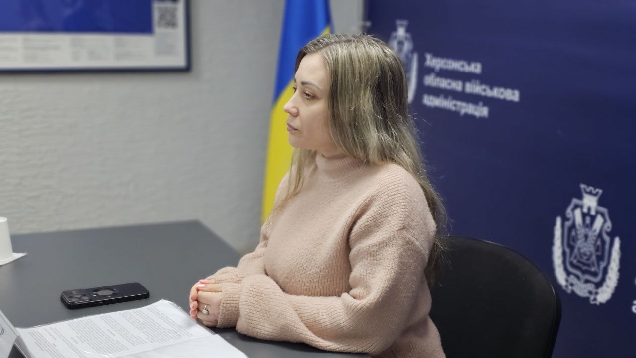Жінка сидить за робочим столом під час наради в офіційній установі. На фоні видно банер із написом «Херсонська обласна військова адміністрація» та державний прапор України.