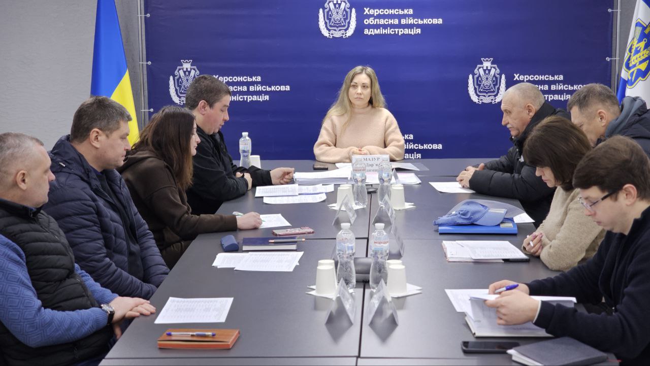 Робоча зустріч у Херсонській обласній військовій адміністрації. Група людей сидить за довгим столом під час обговорення документів. На фоні — синій банер із гербом та офіційною назвою установи, а також прапор України.