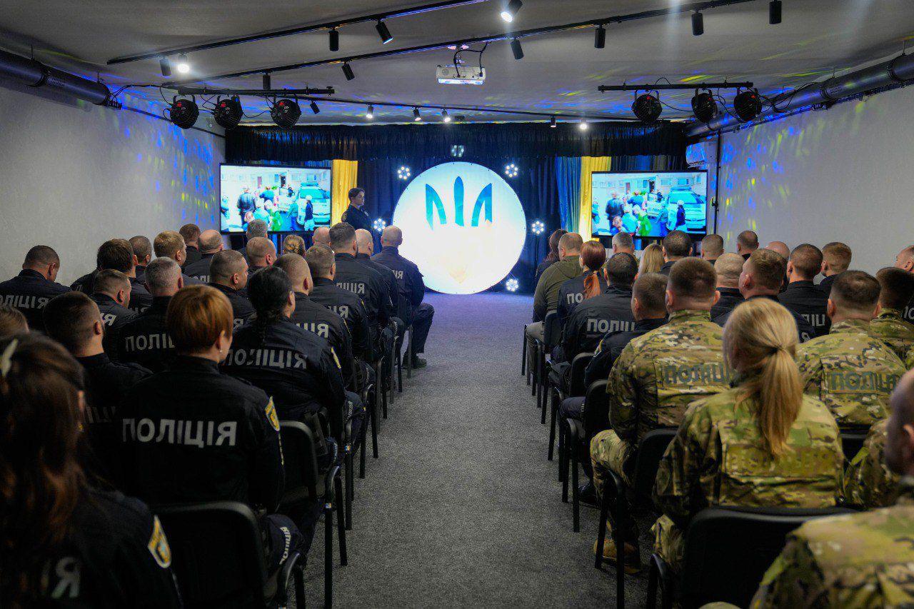 Велика аудиторія, заповнена працівниками поліції та військовими у формі, які сидять спиною до камери та дивляться на сцену. На сцені встановлено круглий світловий екран із логотипом, а з боків розташовані монітори, що транслюють відеоматеріали.