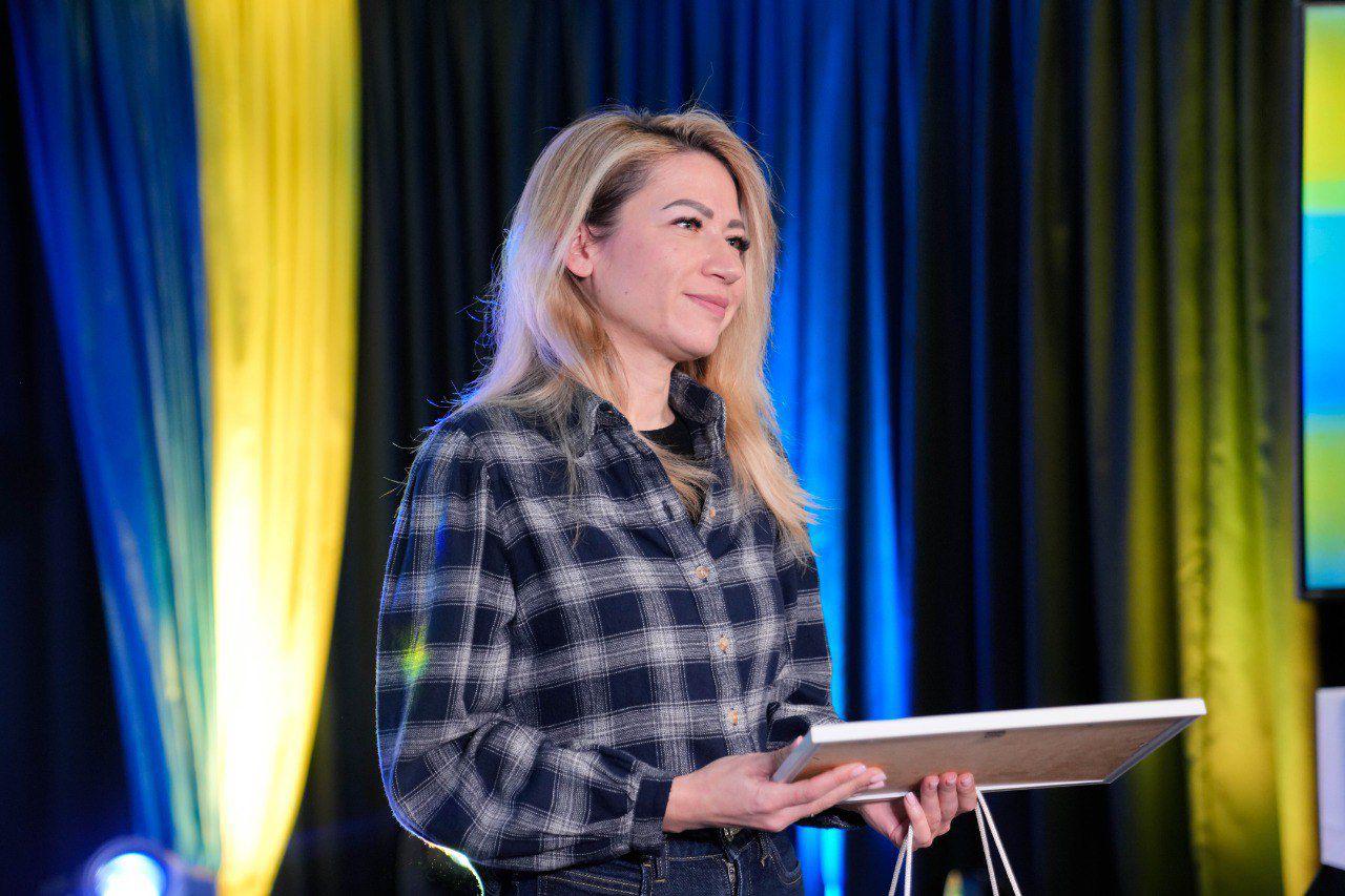 Жінка у картатій сорочці стоїть на сцені та тримає в руках рамку, ймовірно, з дипломом або подякою. На фоні — синьо-жовта завіса, що підсвічується софітами.