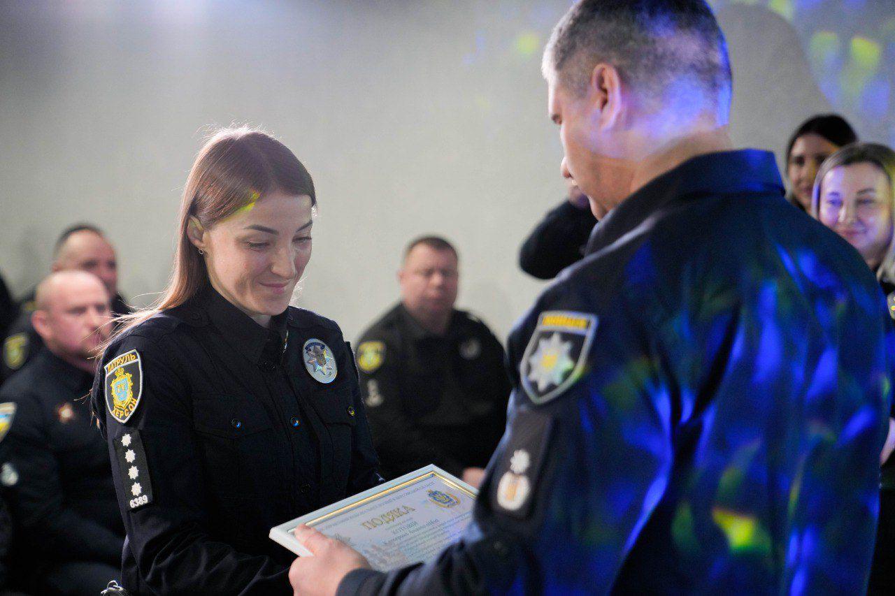 Урочисте нагородження працівниці поліції. Чоловік у формі вручає жінці-офіцеру почесну грамоту (подяку). На задньому плані в залі сидять інші співробітники правоохоронних органів.