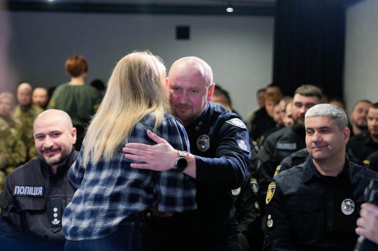 Чоловік у поліцейській формі вітає та обіймає жінку у картатій сорочці під час урочистої церемонії. На задньому плані в залі сидять інші співробітники поліції у формі.