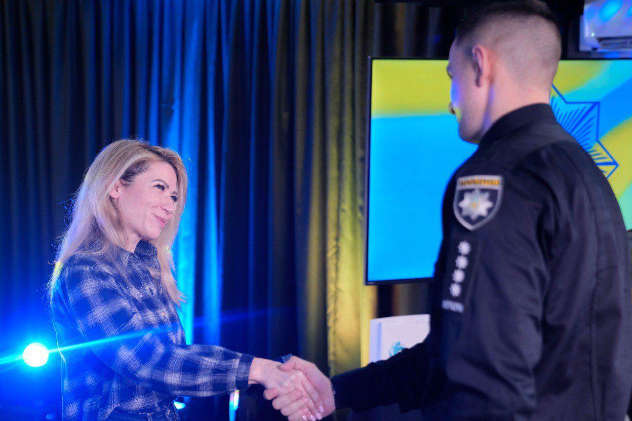 Жінка у картатій сорочці стоїть на сцені та з посмішкою тисне руку чоловікові у поліцейській формі під час офіційної церемонії нагородження. На фоні — синьо-жовта завіса та екран із державною символікою.