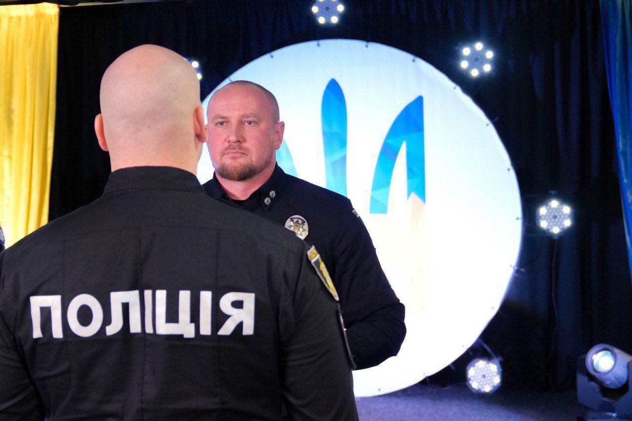 Чоловік у поліцейській формі стоїть на сцені перед великим круглим екраном із логотипом. На передньому плані видно спину іншого співробітника поліції у формі з написом «ПОЛІЦІЯ».