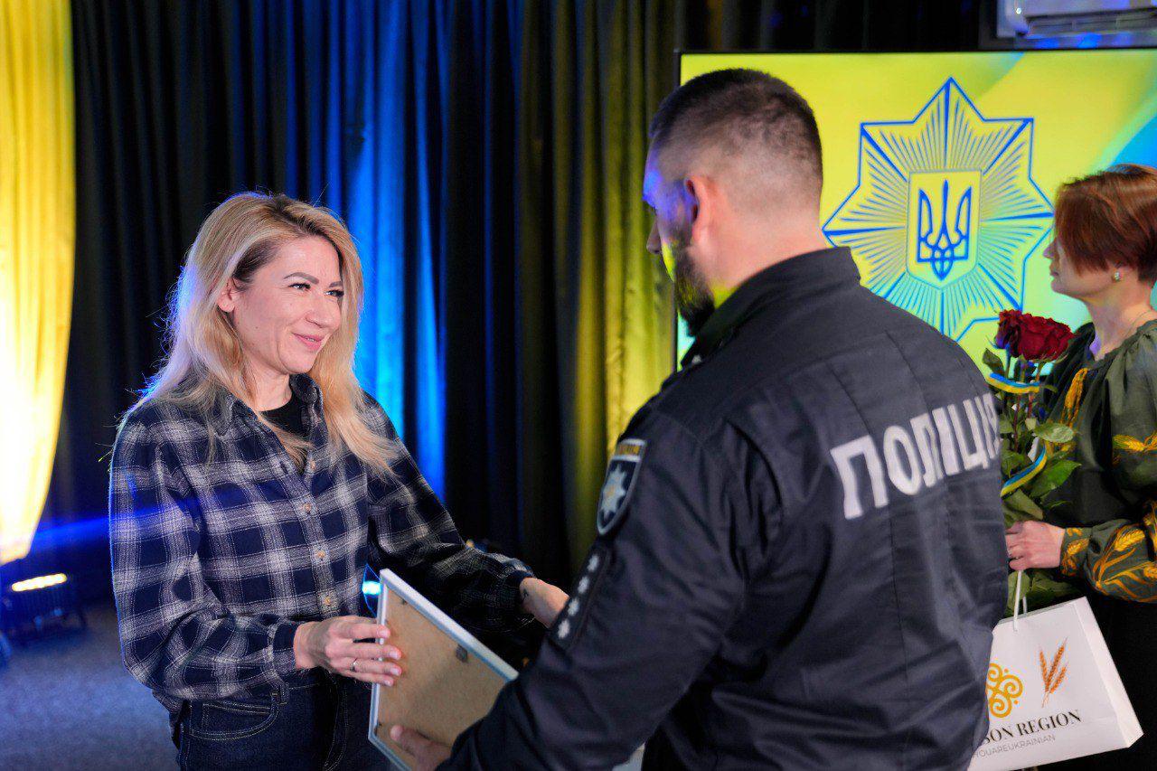 На офіційній церемонії жінка у картатій сорочці вручає нагороду чоловікові у поліцейській формі. Поруч стоїть ще одна жінка з букетом квітів та пакунком із логотипом. На задньому плані — екран із зображенням восьмикутної зірки та герба України.