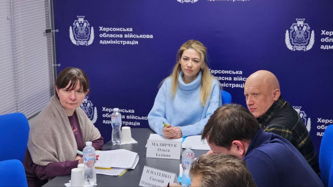 Учасники робочої наради в офіційному приміщенні сидять за столом, обговорюючи робочі питання. Перед ними лежать документи та стоять таблички з іменами. На фоні — темно-синій банер із символікою та написом «Херсонська обласна військова адміністрація».
