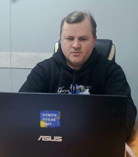 Чоловік у чорному худі працює за ноутбуком. На кришці ноутбука наклеєно стікер із написом «ОСВІТА РУХАЄ СВІТ» на фоні кольорів державного прапора України.