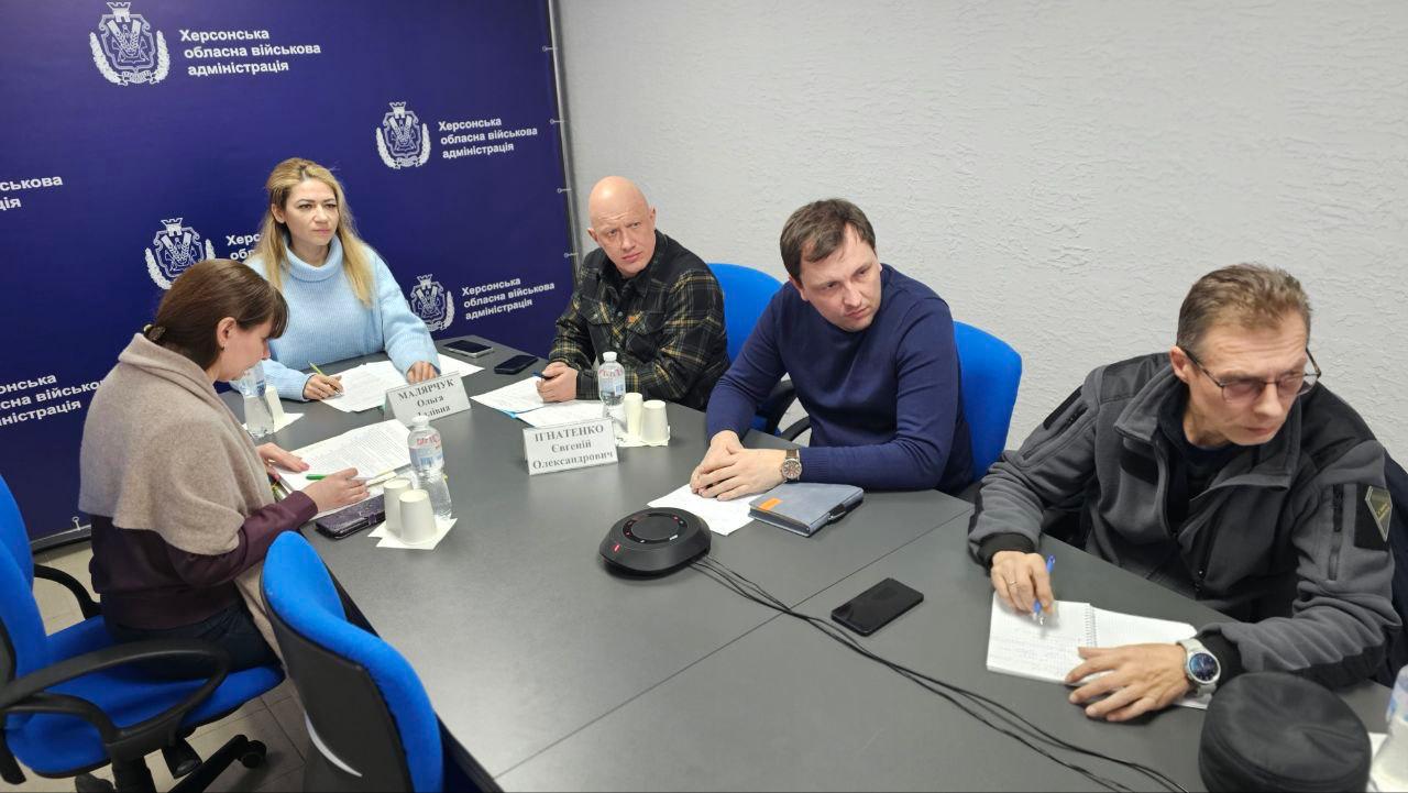 Учасники робочої зустрічі в Херсонській ОВА сидять за столом, обговорюють документи та роблять нотатки. Перед ними розміщено пляшки з водою та таблички з іменами. На фоні — банер синього кольору з офіційною символікою та назвою установи.