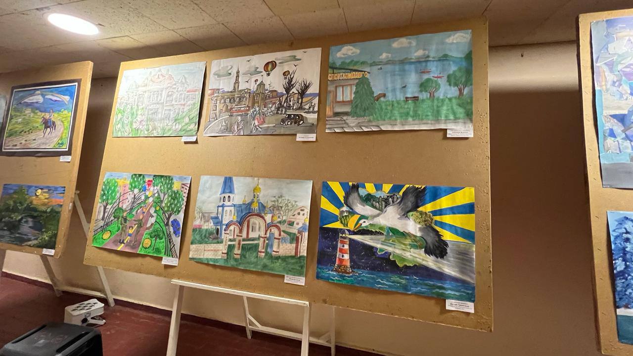 Стенд із дитячими малюнками на виставці. Роботи зображують міські пейзажі, архітектуру з куполами, берег річки та символічний малюнок із лелекою над морем і маяком. Усі малюнки мають етикетки з іменами авторів.