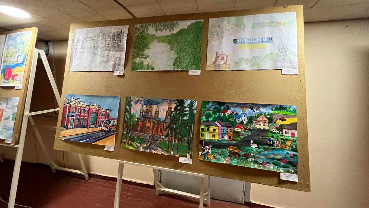 Експозиція дитячих малюнків на великому стенді. Серед робіт — зображення природи, міської архітектури, залізничного вокзалу та патріотичний плакат із написом «Олешки». Кожен малюнок має білу інформаційну картку в нижньому куті.
