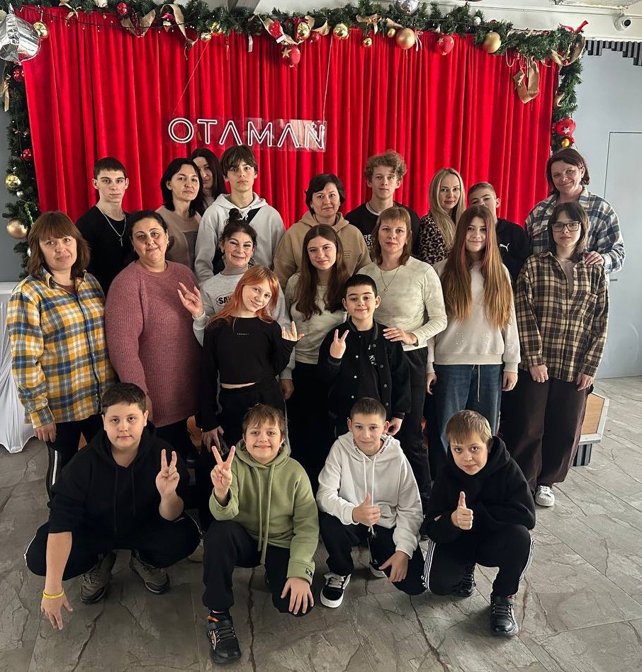 Групове фото дітей та дорослих на фоні червоної завіси з неоновим написом. Люди позують у кілька рядів, деякі показують жести «мир» або «клас». Локація прикрашена різдвяною гірляндою з іграшками.