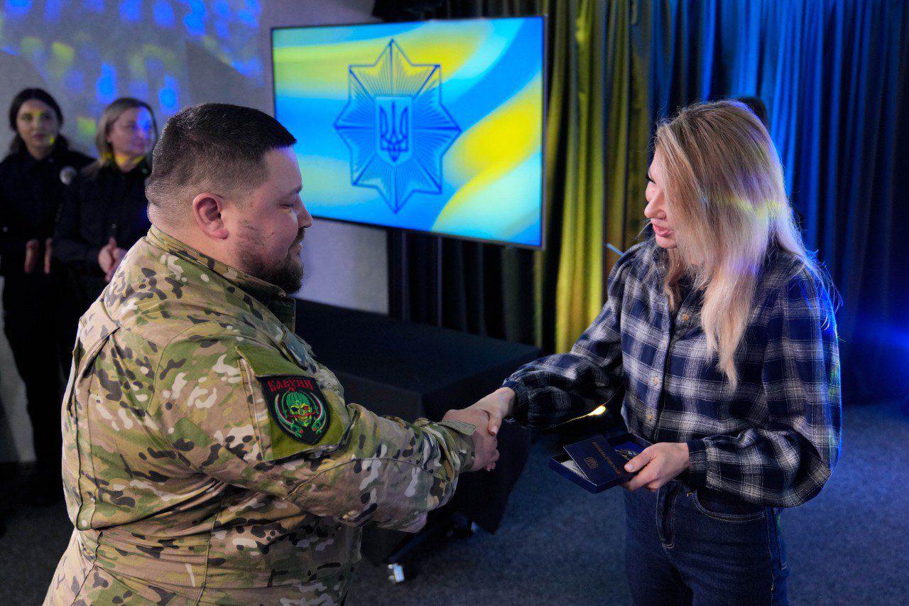 На офіційному заході чоловік у камуфляжній формі тисне руку жінці в картатій сорочці, яка тримає в руках нагороду. На задньому плані встановлено екран із символікою Національної поліції України, а поруч стоять інші співробітники у формі.