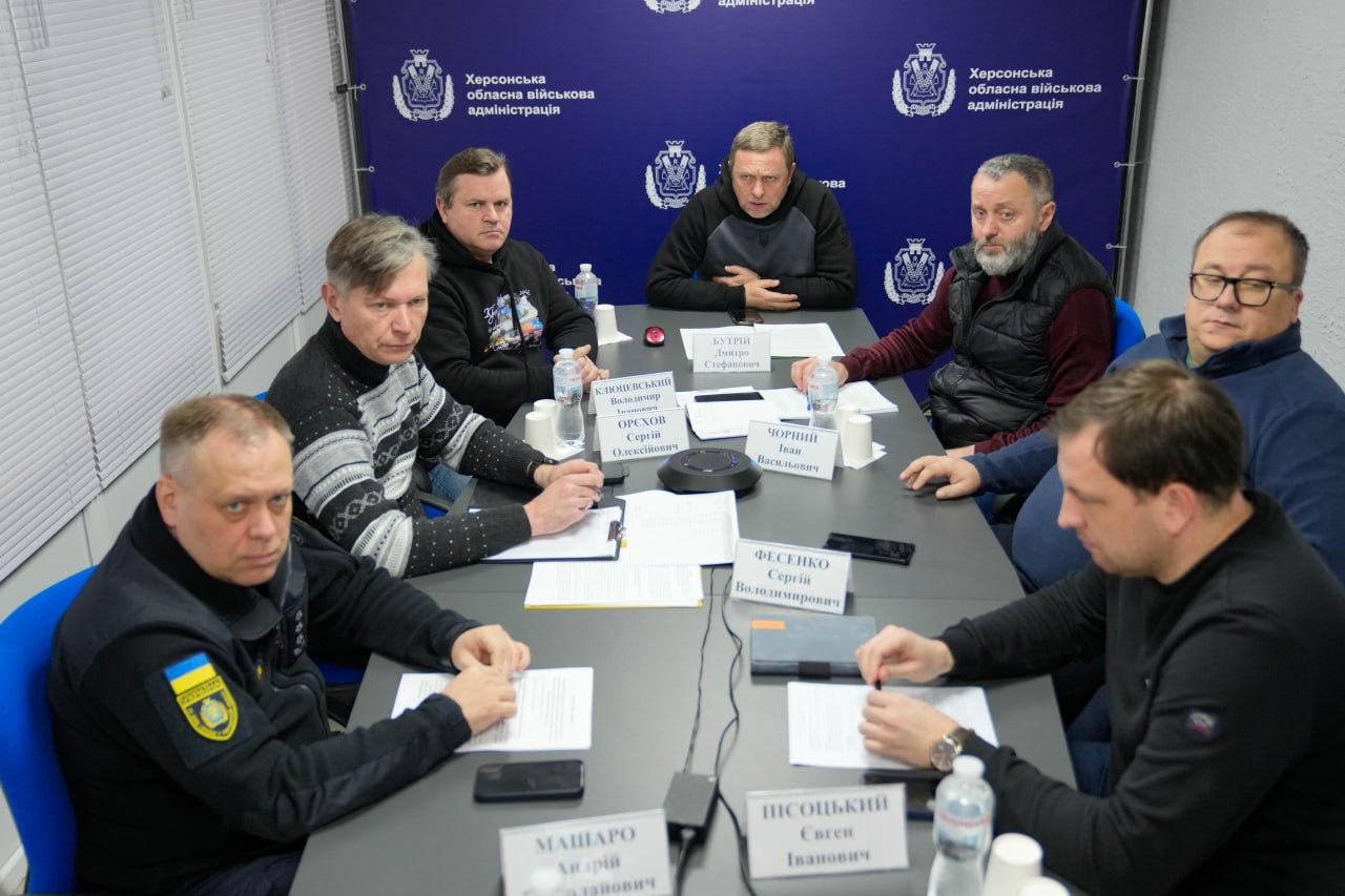 Група чоловіків проводить робочу зустріч за великим столом у кабінеті. Перед учасниками розкладені документи, блокноти та таблички з іменами. На задньому плані — синій банер із гербом та написом «Херсонська обласна військова адміністрація».