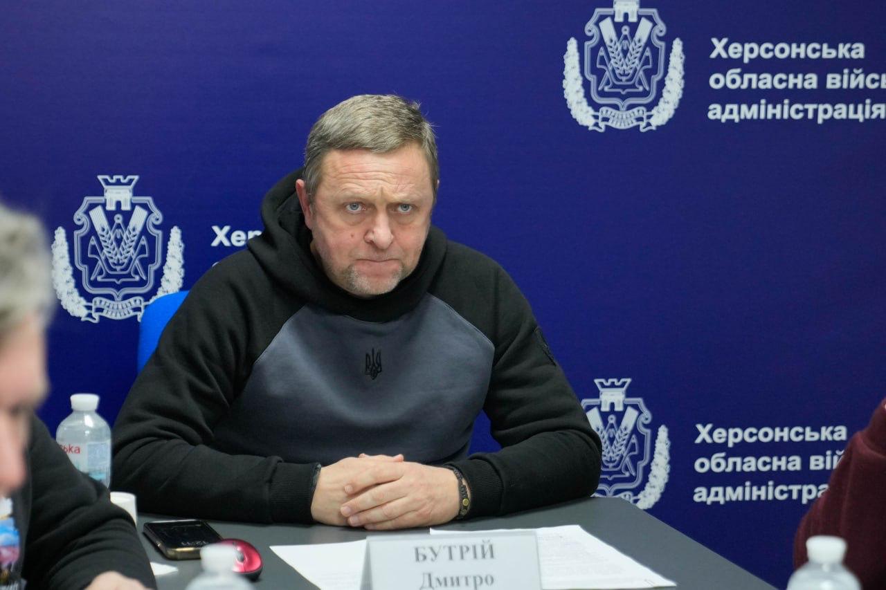 Чоловік у чорному худі з невеликим гербом України сидить за столом під час офіційної зустрічі. Він тримає руки зчепленими перед собою. На фоні за ним — синій банер із символікою та написом «Херсонська обласна військова адміністрація».