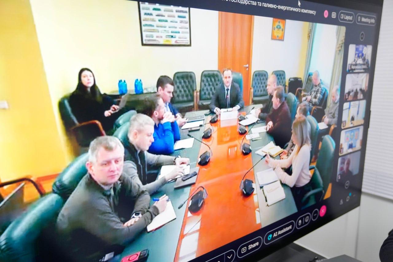 Фотографія екрана, на якому відображено онлайн-нараду. Учасники засідання сидять за довгим столом у конференц-залі, а збоку на моніторі видно вікна з іншими підключеними користувачами. На столі розміщені мікрофони, ноутбуки та папери.