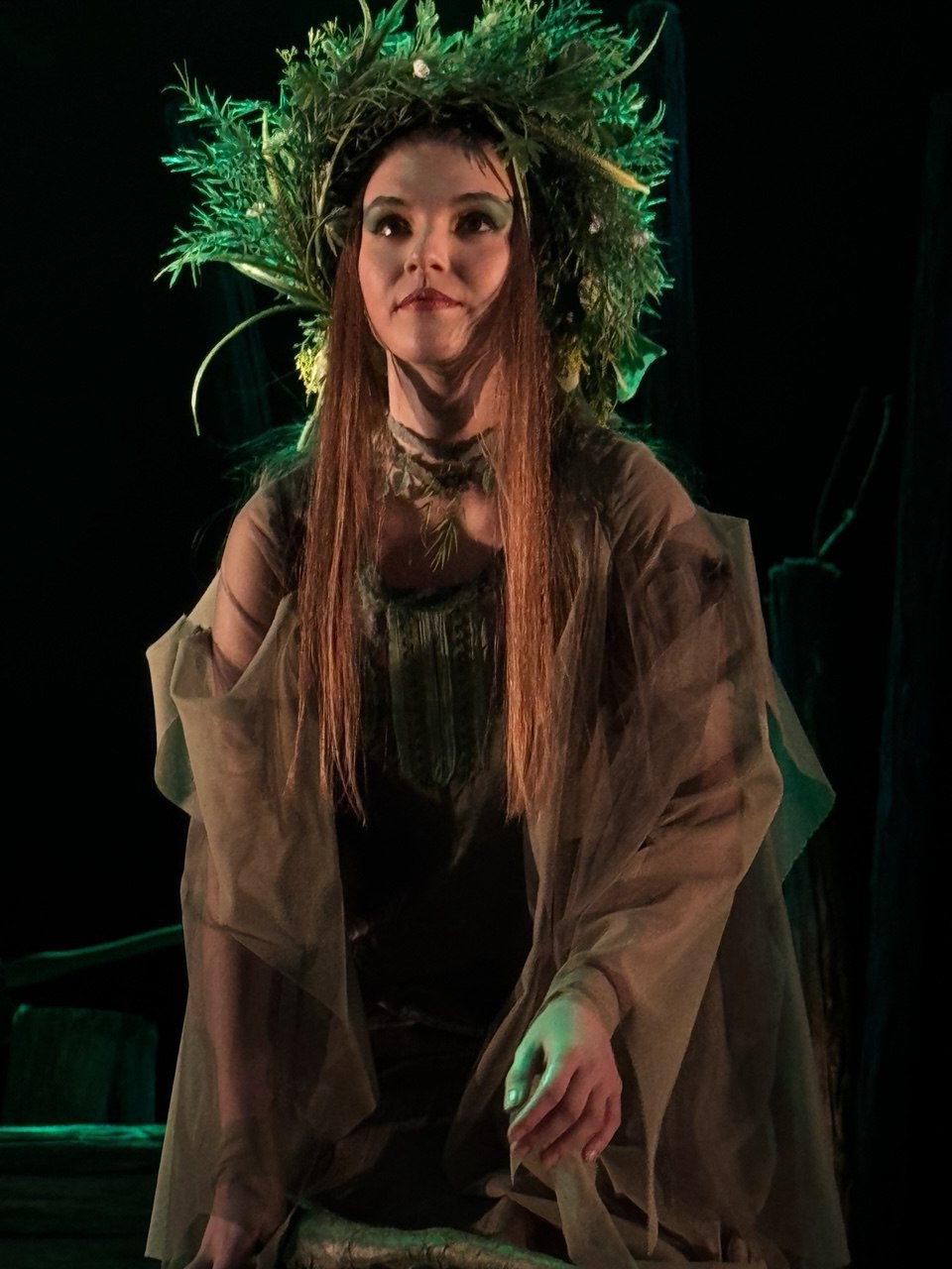 Акторка в образі міфічної лісової істоти на театральній сцені. Вона одягнена в легке вбрання зелених і коричневих відтінків, а на голові має об'ємний вінок із густої зелені та гілок. Погляд спрямований вгору, освітлення сцени створює таємничу атмосферу.