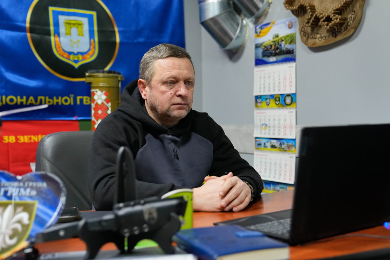 Чоловік у чорному худі сидить за робочим столом перед відкритим ноутбуком. На столі лежать документи та канцелярія. На стіні за ним розміщено календар, українську символіку та прапор із гербом.