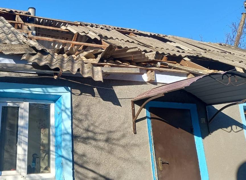 Фасад одноповерхового будинку з пошкодженим шиферним дахом. На покрівлі видно великі дірки та зміщені листи шиферу, що відкривають дерев'яну обрешітку. Стіни будинку сірого кольору, вікна та двері мають блакитне обрамлення.