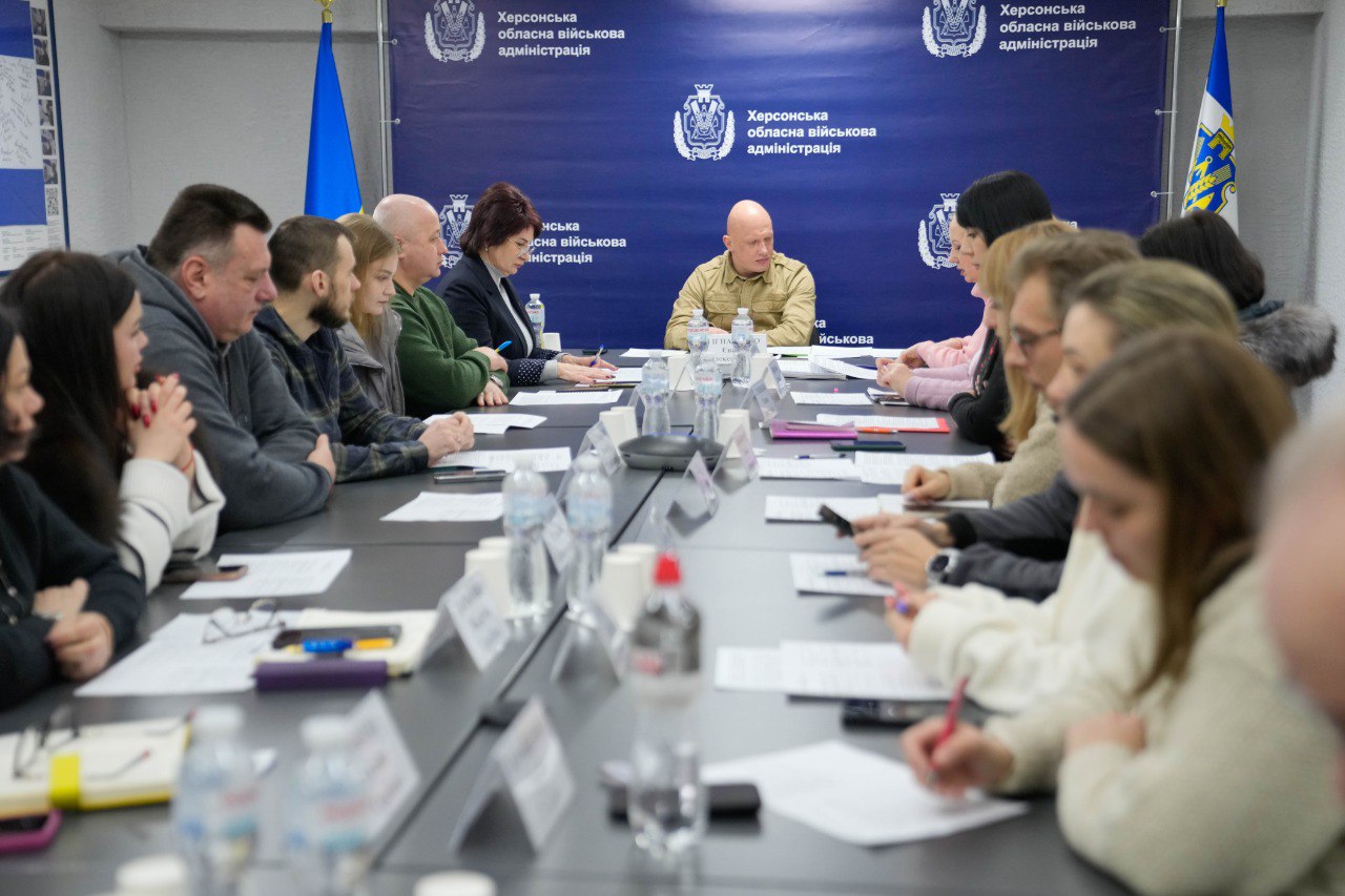Навколо довгого столу в офісному приміщенні проходить робоча зустріч або засідання. За столом сидять чоловіки та жінки з паперами та блокнотами, а в центрі, на фоні синього банера з офіційною символікою Херсонської обласної військової адміністрації, сидить керівник.