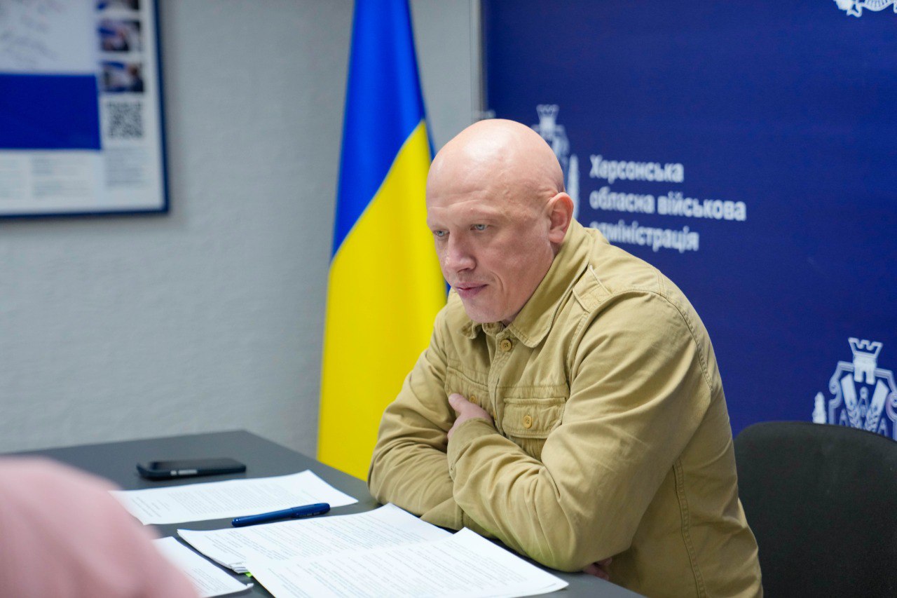 Чоловік у сорочці пісочного кольору зосереджено працює з документами за столом на фоні прапора України та офіційного банера.