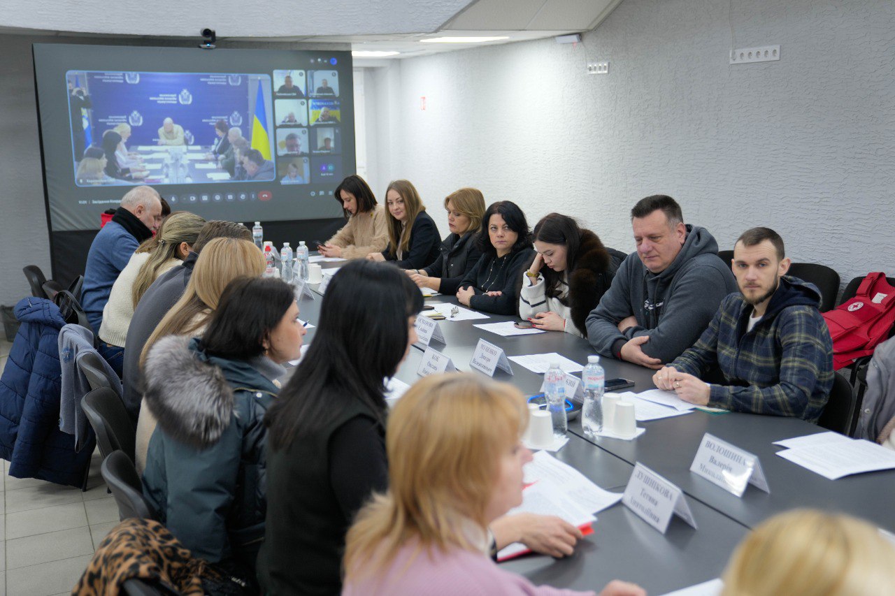 Робоча нарада у просторому кабінеті. Учасники сидять за довгим столом, а на великому екрані транслюється відеоконференція з іншими колегами.