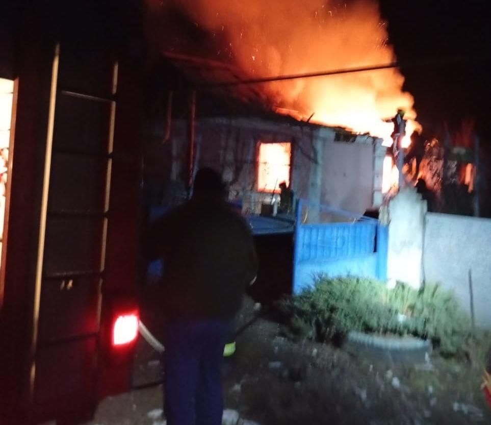 Нічний кадр пожежі в житловому будинку. Крізь вікна та дах будівлі видно яскраве полум'я та густий дим. На передньому плані помітно силует рятувальника та частину пожежного автомобіля з увімкненим ліхтарем.