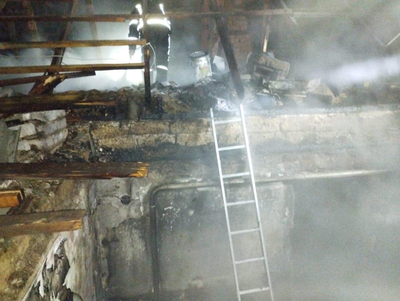 Задимлене приміщення будівлі після пожежі. У центрі стоїть металева драбина, що веде до зруйнованого перекриття. На верхньому рівні крізь густий дим видно силует рятувальника у світловідбивній формі, який оглядає пошкоджені конструкції.