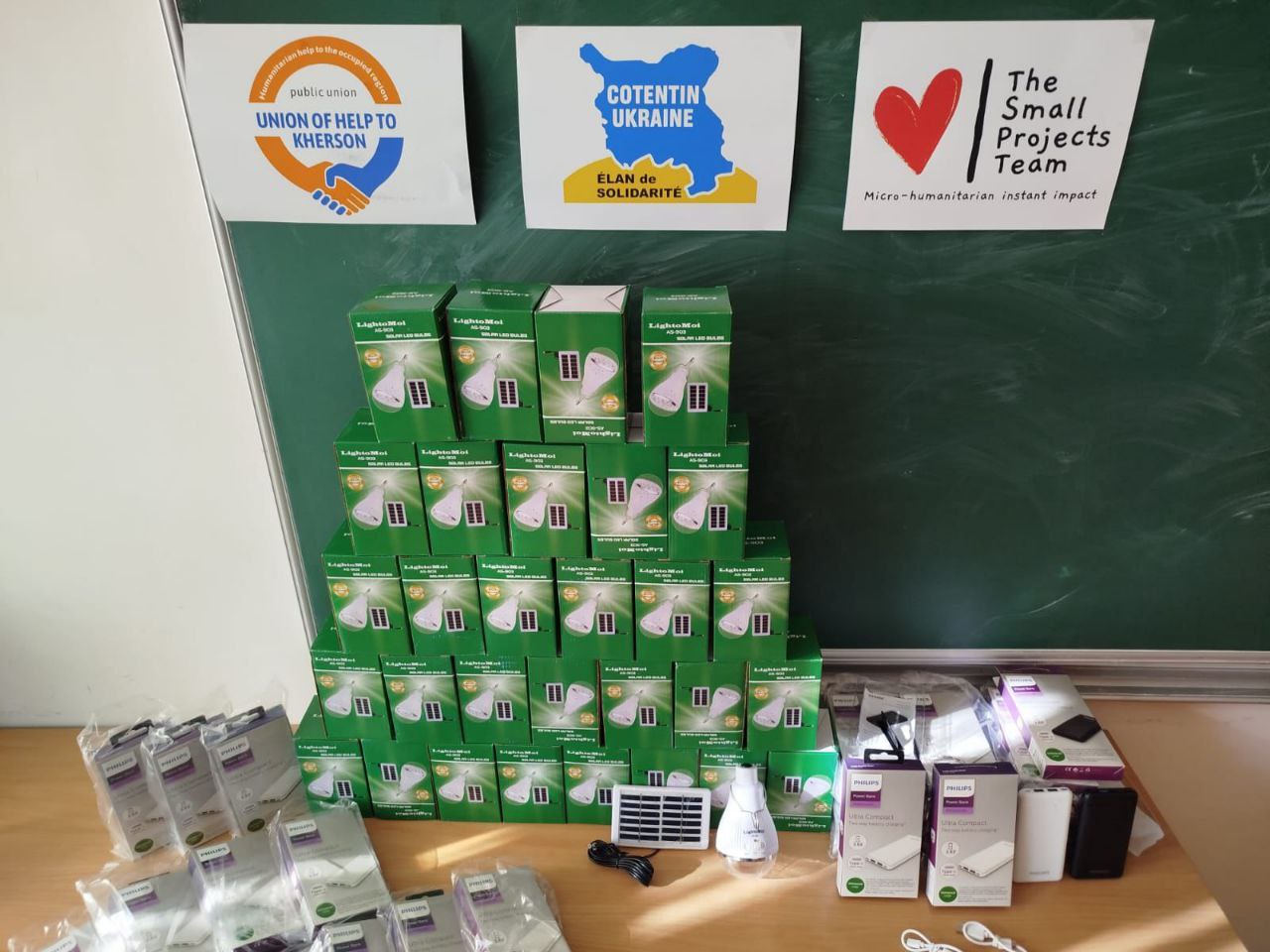 Гуманітарна допомога на столі перед шкільною дошкою. На задньому плані розміщено плакати благодійних організацій: «Union of Help to Kherson», «Cotentin Ukraine» та «The Small Projects Team». На столі складено пірамідою численні коробки з LED-лампами на сонячних батареях, а поруч — портативні зарядні пристрої (павербанки) Philips.