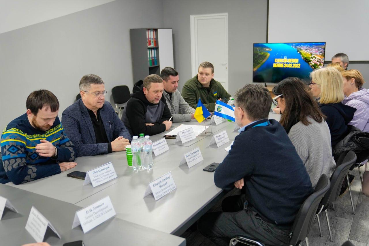 Група людей проводить офіційну зустріч за довгим столом у конференц-залі. На столі розміщені таблички з іменами, пляшки з водою та прапори України й Херсонської області. На фоні видно телевізор із заставкою про Херсонщину та стелаж із папками.