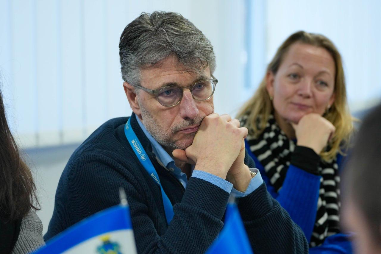 Портрет учасника офіційної зустрічі — чоловіка в окулярах та синьому светрі з брендованою стрічкою UNHCR (УВКБ ООН) на шиї. Він зосереджено слухає, спершись підборіддям на руки. Поруч на фоні видно іншу учасницю зустрічі.